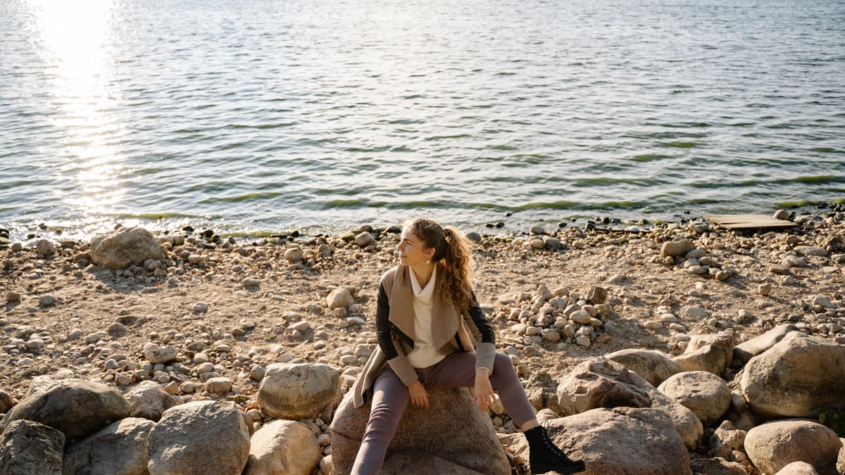 A woman sits on a rocky shoreline, gazing at the serene lake view during a sunny day.